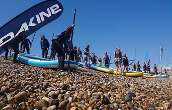 People preparing large paddleboards on a pebbled beach under a clear blue sky.