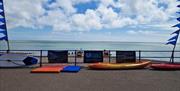 Bourne to Kayak banners fixed to blue railings with the beach, sea and blue sky in the background, with a kayak and stand up paddle board displayed in
