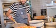 Chef in a striped t-shirt and grey apron using a pizza paddle to transfer a pizza onto a plate from the oven