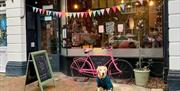 Black restaurant exterior with a pink bicycle leant against the wall and multi coloured bunting in the window. A blackboard is displayed outside of th