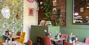 Interior of The Good Grub restaurant with green leaf patterned wallpaper, exposed bricks and wooden tables with yellow and pink chairs.