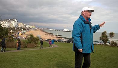 Walking guide in a blue waterproof coat and white baseball cap standing on the grassy wish tower slopes with a view of the seafront and pier in the ba