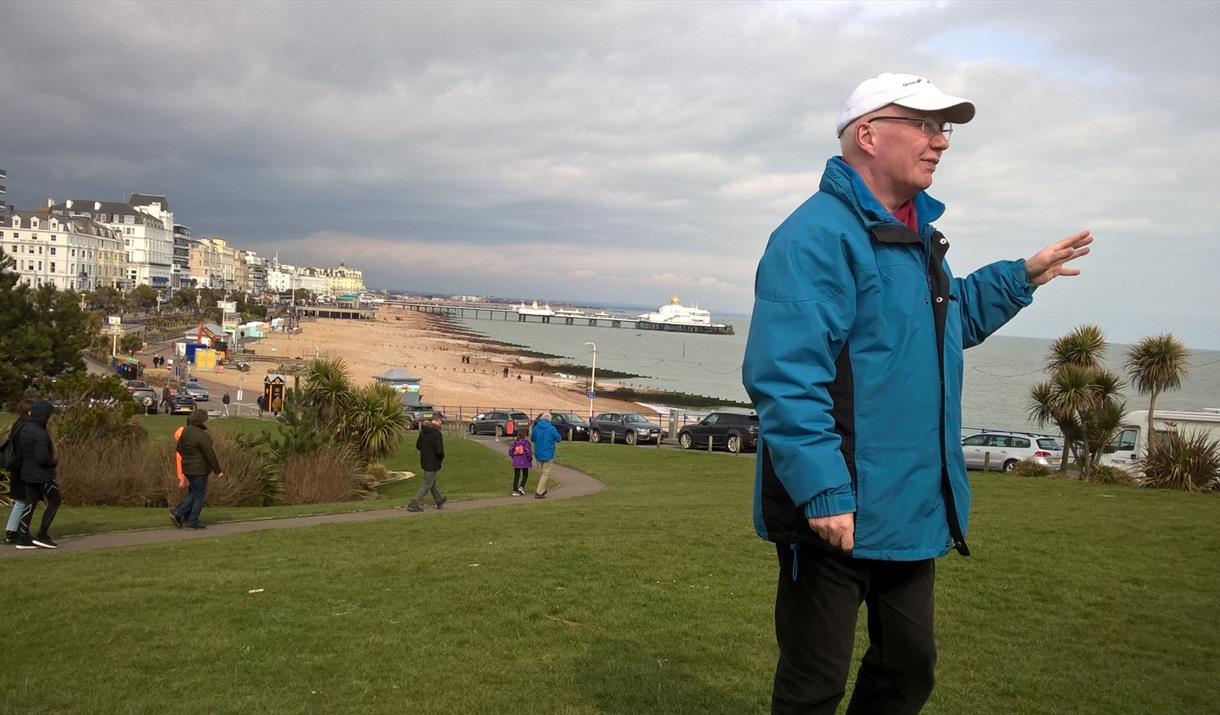 Walking guide in a blue waterproof coat and white baseball cap standing on the grassy wish tower slopes with a view of the seafront and pier in the ba