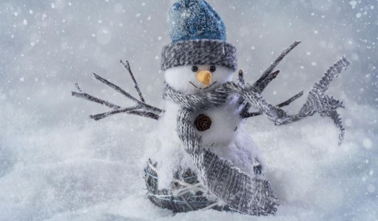 Snowman craft made from white wool pom poms wearing a blue hat with stick arms at either side. Standing in a snowy scene