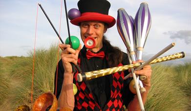 A circus performer holding lots of equipment in a comedic way including juggling batons and diablos.