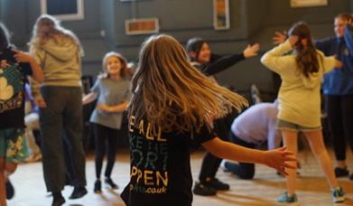 A room of children and young people playing and performing with a girl spinning in the foreground wearing a Doorstep Arts tshirt