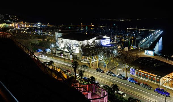 A view of the whole theatre from rock walk steps with the marina in the background. the venue is lit white at night time.