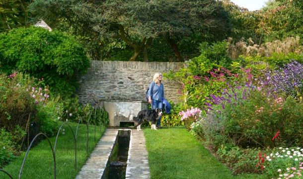 A woman walking through the formal gardens at Coleton Fishacre. The image is framed by blooming flowers beds, and a structured stream carves through t
