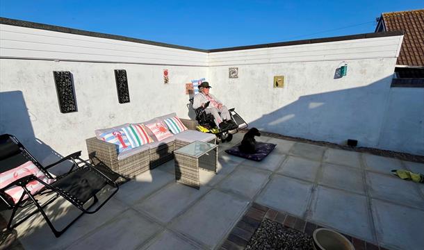 Back garden, with patio furniture and sun loungers. Wheelchair access through side gate from driveway. A person is in a wheelchair with a black Giant
