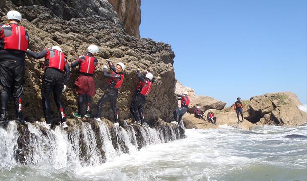 Participants in a coasteering session traverse a rocky wall between coasteering jumps. The participants are wearing wetsuits, buoyancy aids and helmet