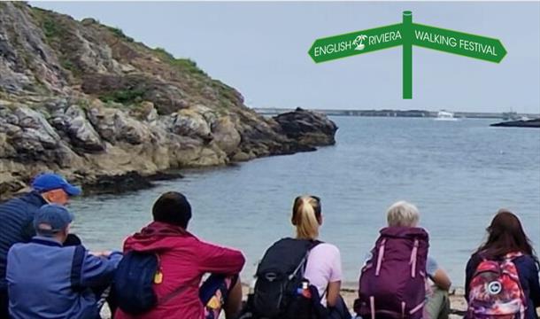 A group of people in walking clothes sitting side by side looking out across the water at Churston Cove, Brixham
