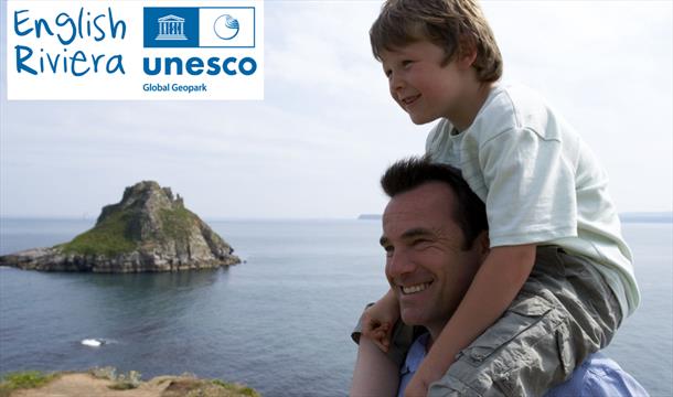 A man with a young boy sitting on his shoulders,  standing on the South West Coast Path with Thatchers Rock, Torquay, and the sea in the background. E