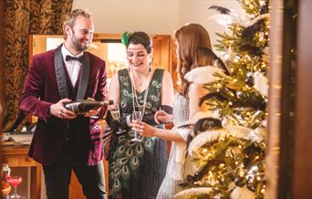 Three people dressed in vintage 1920s clothing drink champagne by a gold and black Christmas tree