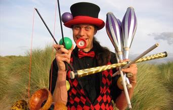 A circus performer holding lots of equipment in a comedic way including juggling batons and diablos.