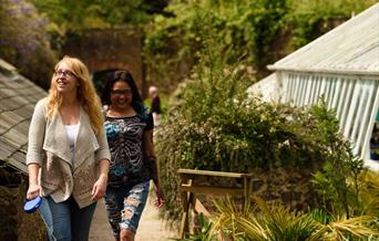 Two women walking through the formal gardens at Greenway. The image is framed by blooming flowers beds, and a glimpse of the wooden framed greenhouse