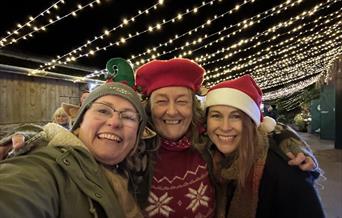 Three ladies in Santa hats enjoying the festive lights in the Stable Yard at Cockington Court Craft Centre