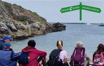 A group of people in walking clothes sitting side by side looking out across the water at Churston Cove, Brixham