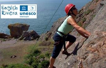 A rock climber on rock face, with sea in background. English Riviera UNESCO Global  Geopark.