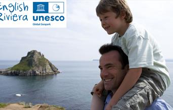 A man with a young boy sitting on his shoulders,  standing on the South West Coast Path with Thatchers Rock, Torquay, and the sea in the background. E