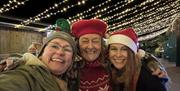 Three ladies in Santa hats enjoying the festive lights in the Stable Yard at Cockington Court Craft Centre