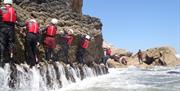 Participants in a coasteering session traverse a rocky wall between coasteering jumps. The participants are wearing wetsuits, buoyancy aids and helmet