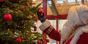 Santa Claus stood next to a Christmas tree, holding a bauble