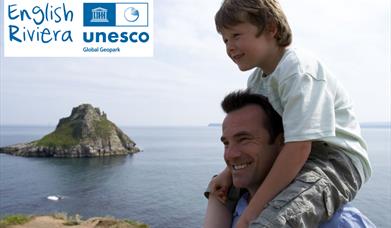 A man with a young boy sitting on his shoulders,  standing on the South West Coast Path with Thatchers Rock, Torquay, and the sea in the background. E