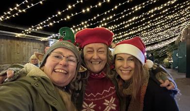 Three ladies in Santa hats enjoying the festive lights in the Stable Yard at Cockington Court Craft Centre