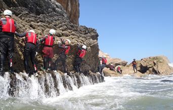 Participants in a coasteering session traverse a rocky wall between coasteering jumps. The participants are wearing wetsuits, buoyancy aids and helmet