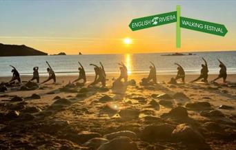 A group of people doing a yoga stretch at sunrise on the beach, with the sea in the background.