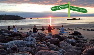 A meditation session with a group of people sitting on the beach at sunset, facing the sea.