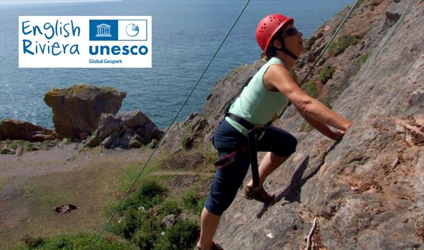 A rock climber on rock face, with sea in background. English Riviera UNESCO Global  Geopark.