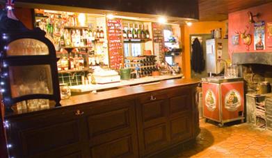 A photo of the bar area at Olivers Jazz Bar, The Bar is wooden and behind it is shelfs filled with bottles. To the right of the bar is a small old fa