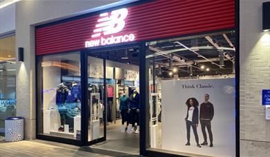 Outside New Balance at The O2. A stylish shopfront with a red banner and windows showing off all their great products.