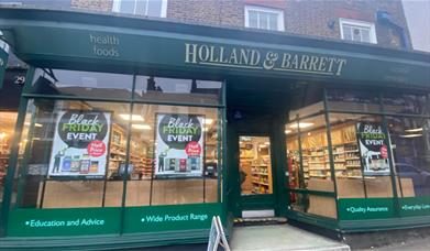 Outside Holland & Barrett in Woolwich. Showing a green shop-front with glass windows and posters.