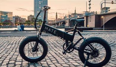 A picture of a black and white mate bike, set next to a river and infront of a stunning sunset.