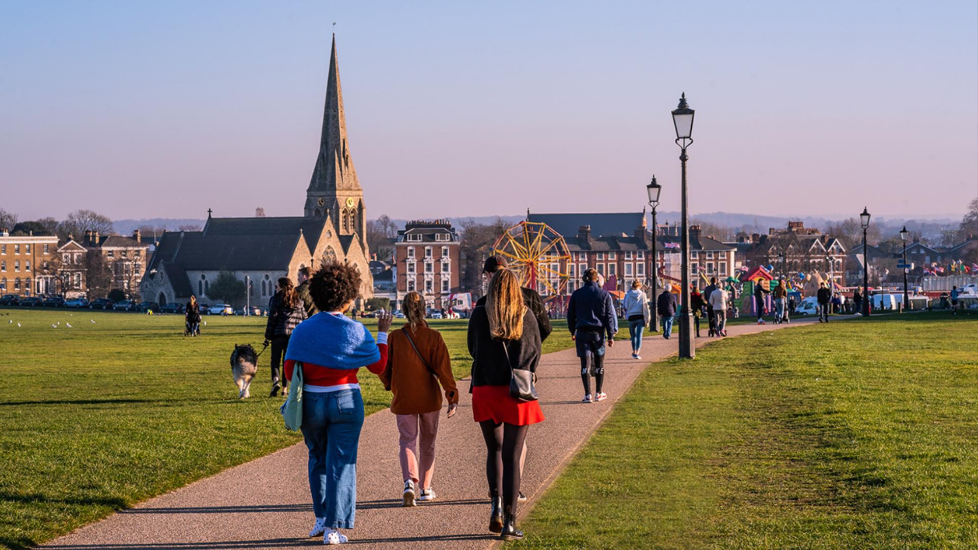 People walk across Blackheath towards All Saints Church and a fairground.