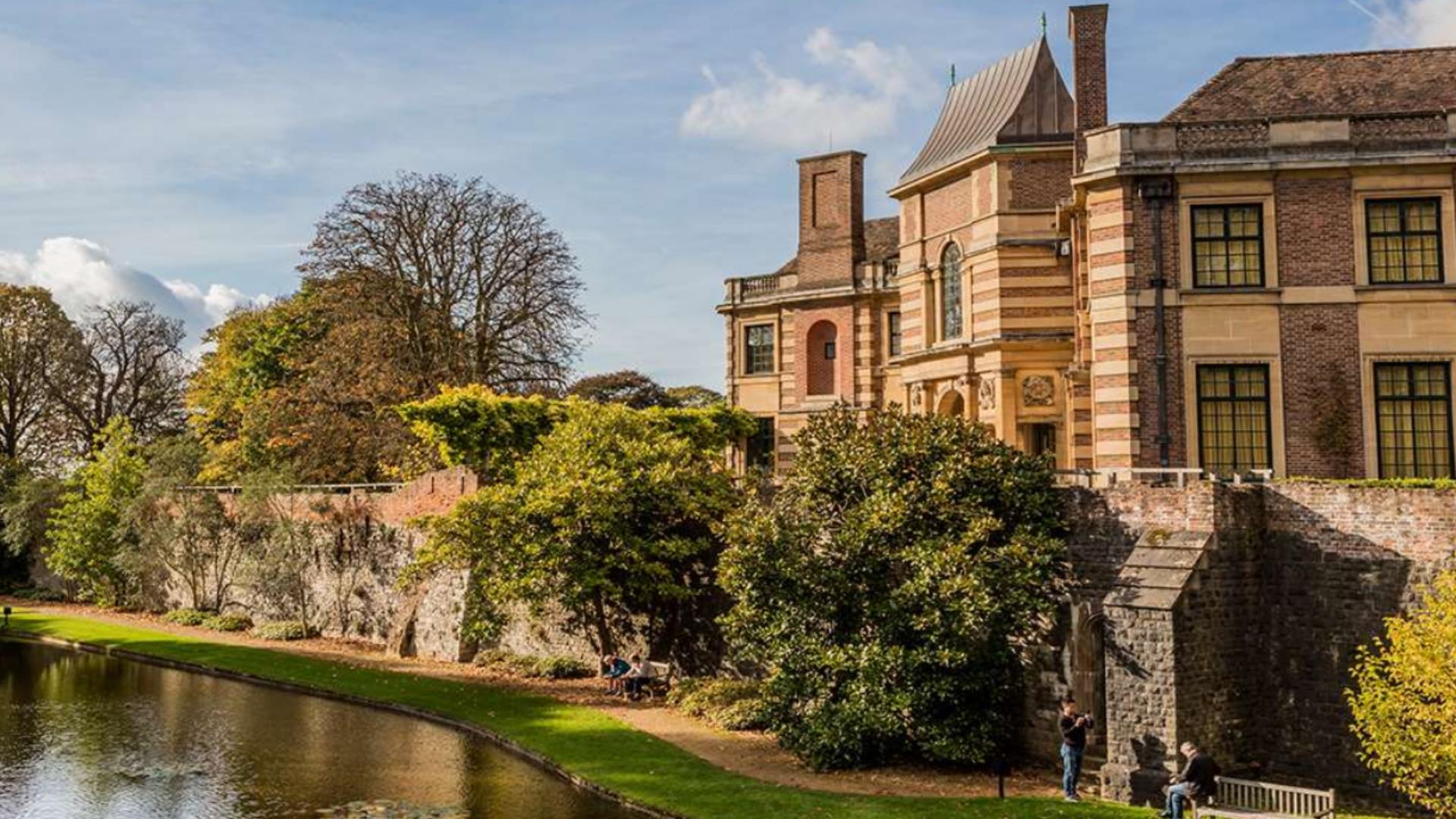 A couple sit outside Eltham Palace and Gardens beside its moat in the sun.