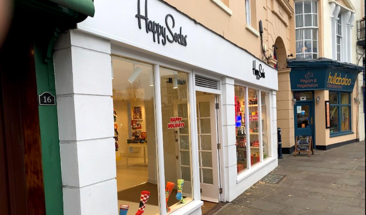 Outside Happy Socks in Greenwich. A white shop front with a black happy socks logo. Inside the windows you can see a unique selection of socks on disp