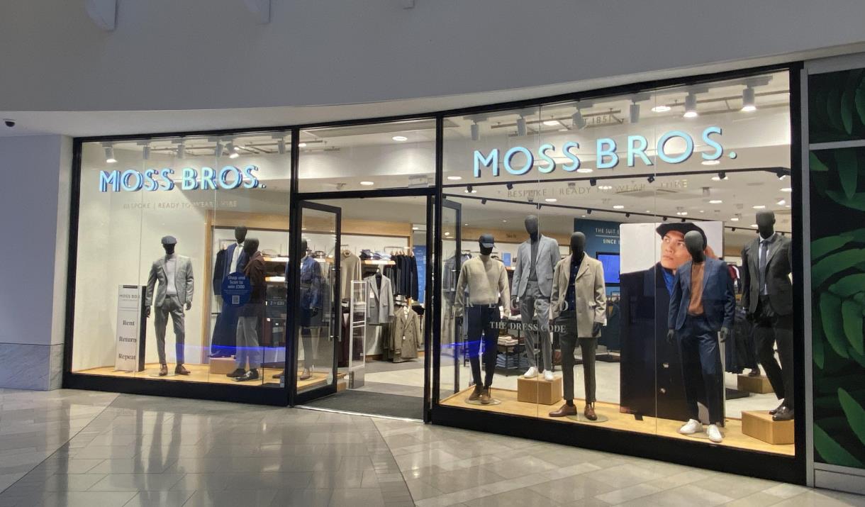 Outside Moss Bros at The O2. A modern shop with a brilliant window display, showing multiple mannequins wearing suits.