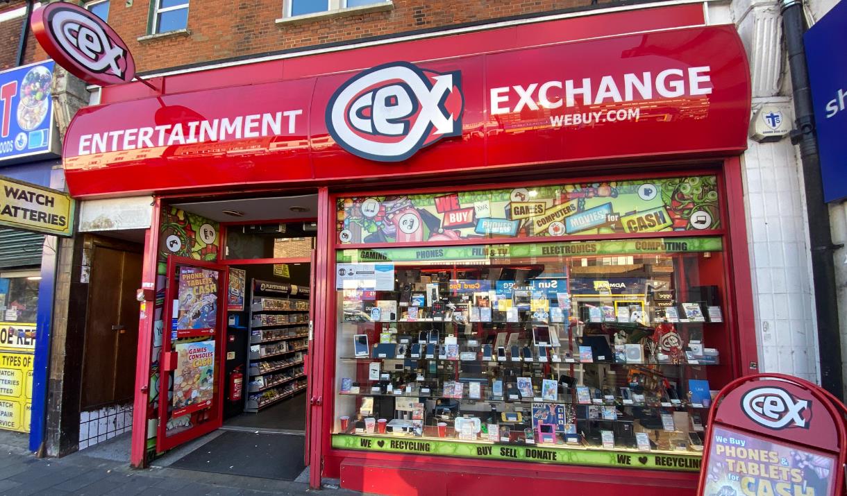 Outside CeX in Eltham. Showing a red and white shop with a selection of electronics presented through the window.