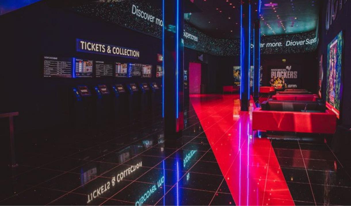 A well taken photo of inside of Cineworld O2, in the picure you can see the tickets and collection area, red and black sofas, movie posters and the en