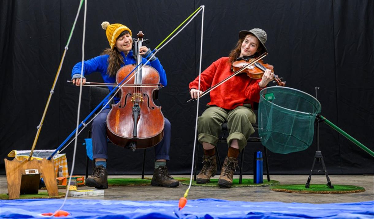 Two musicians settle beside a shimmering lake, fishing into the water.