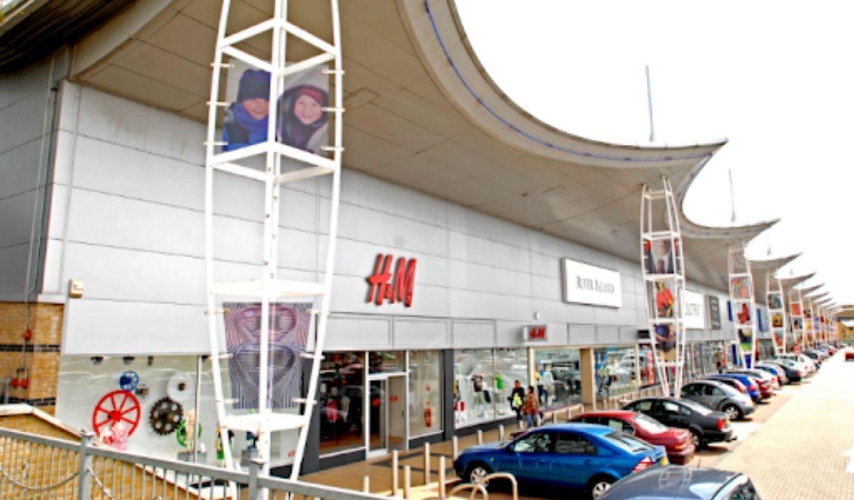 A picture of Greenwich Shopping park, showing a long row of shops. Right in front of them is the car park for easy access.