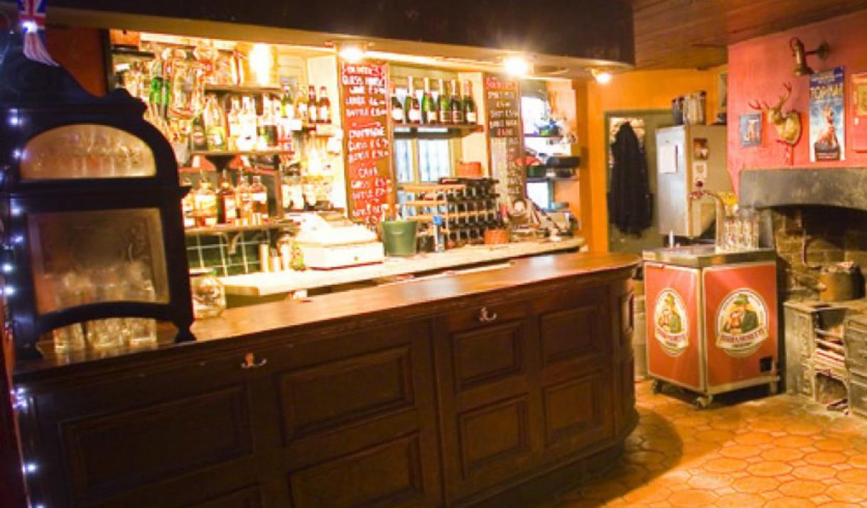 A photo of the bar area at Olivers Jazz Bar, The Bar is wooden and behind it is shelfs filled with bottles. To the right of the bar is a small old fa