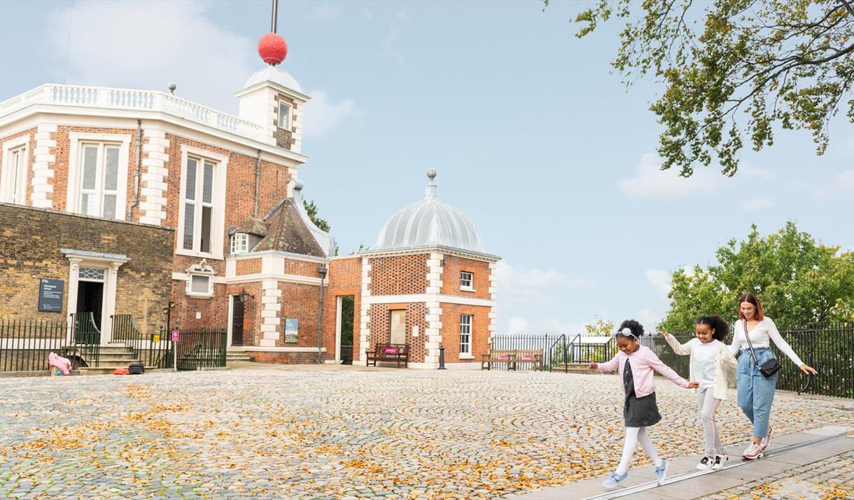 A photo outside of the Royal observatory entrance, there is blue skies, trees and a blanket of orange leaves spread across the stone pavement. Too the