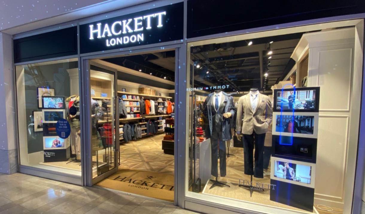 Outside Hackett inside The O2. A modern and welcoming shop front with open doors and windows presenting a range of clothing.