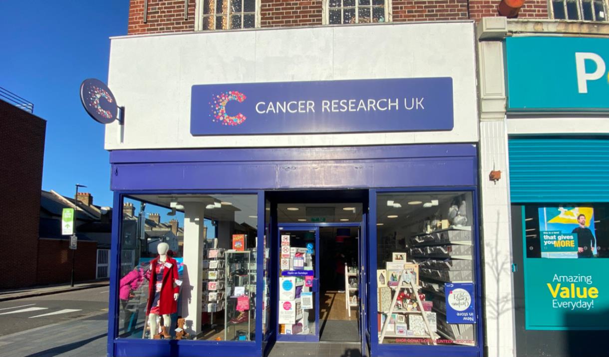 Outside Cancer Research UK, Eltham. A blue and white shop with a cancer research banner at the top. In the windows is a huge selection of items availa