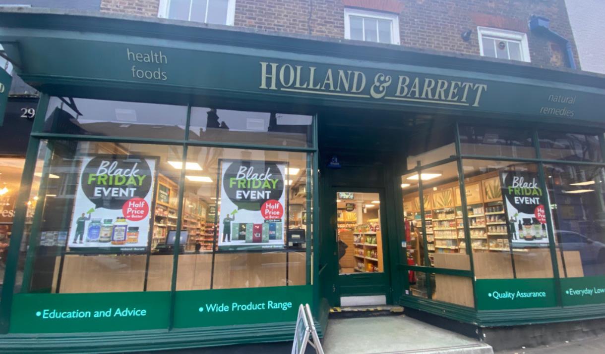 Outside Holland & Barrett in Woolwich. Showing a green shop-front with glass windows and posters.