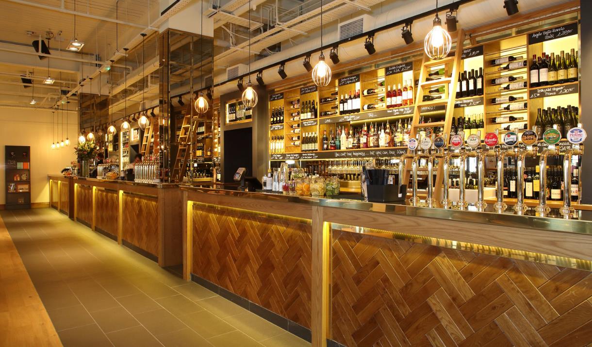 All-Bar-One bar image with well lit up counter with a range of beer taps and a wide selection of wine bottles on the shelves behind.